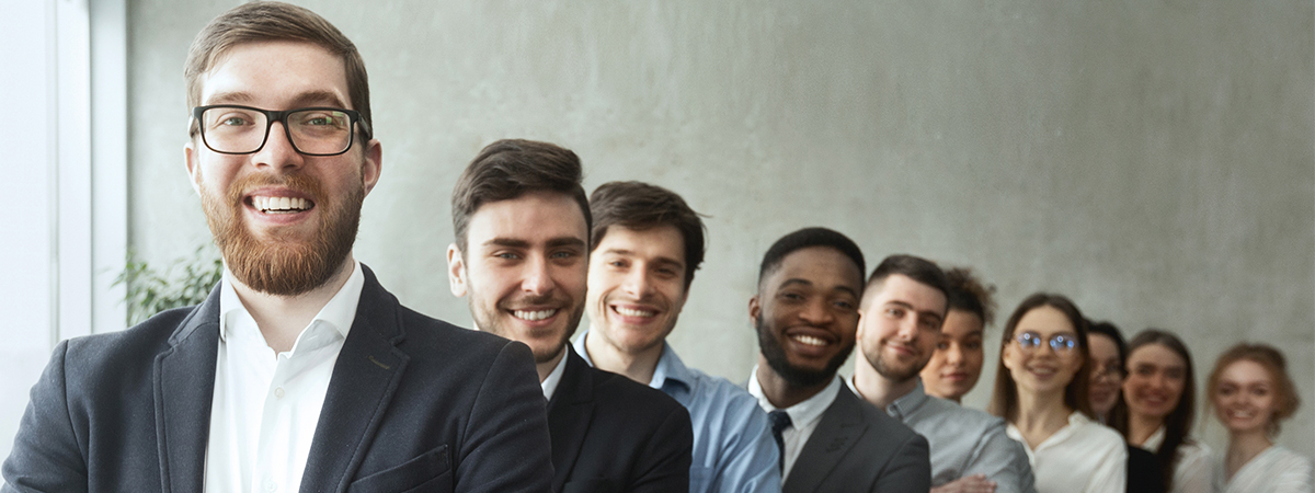 Eine Gruppe von zehn jungen, beruflich gekleideten Menschen steht lächelnd in einer Reihe. Die Person ganz vorne, ein Mann mit Brille und Bart, blickt direkt in die Kamera. Die Gruppe steht in einem modernen Büro vor einer grauen Wand.